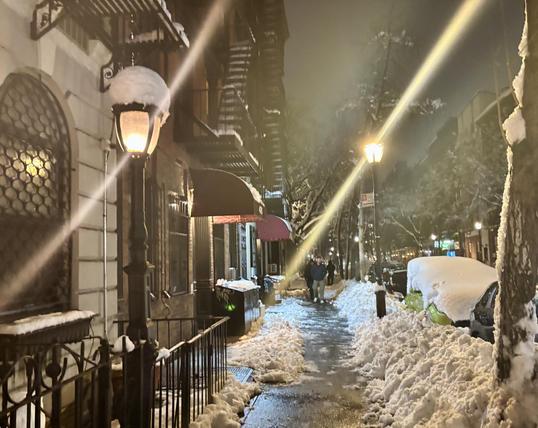 NYC side street sidewalk, skinny path shoveled for walking. Street lamps twinkle.