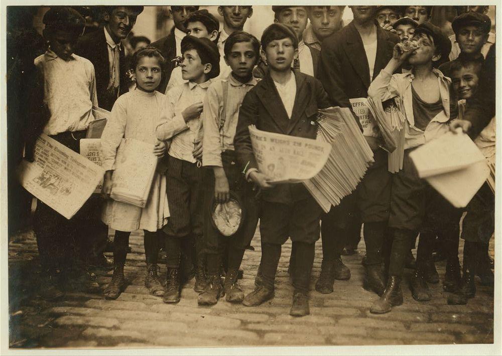 Photo from 1910 of children in a couple of rows, many holding newspapers.