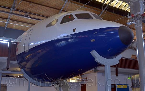 Close up of the nose section of a partial fuselage on display inside a museum.
The plane is white on the top and dark blue underneath, with no other markings visible.
Curved grey metal supports on tall poles hold the front and rear of the fuselage section, the front one almost making it look like the plane is smiling.
Grey walls can be seen under the plane, with more grey walls behind a brown wooden balustrade on the upper level in the background, level with the planes.
A wooden ceiling with metal support beams, and glazed panels in the roof, fill the rest of the frame.
