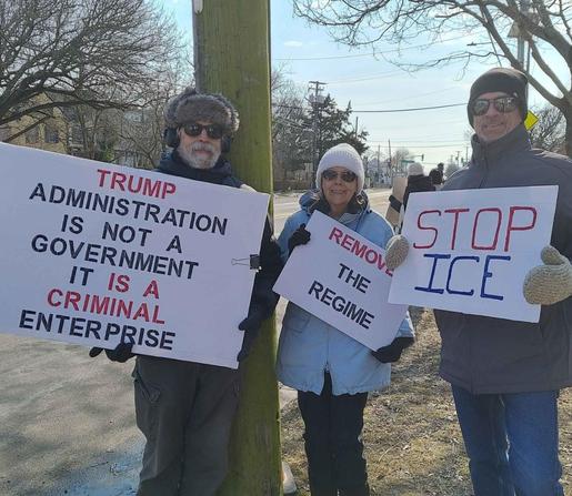 Two men and a woman are standing outside holding handmade protest signs.  They are dressed in warm winter clothing.  The man on the left is holding a sign that reads, "TRUMP ADMINISTRATION IS NOT A GOVERNMENT.  IT IS A CRIMINAL ENTERPRISE." The woman in the middle is holding a sign that says, "REMOVE THE REGIME." The man on the right has a sign that reads, "STOP ICE."