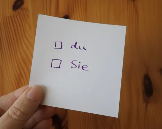 Eine Hand hält einen Zettel, auf dem zum Ankreuzen die beiden Optionen du oder Sie stehen