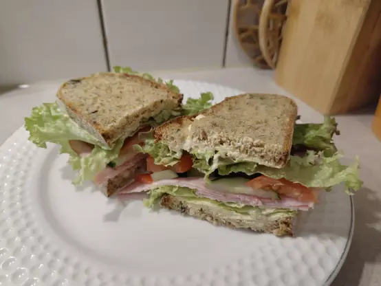 Ein halbiertes Sandwich üppig belegt mit Salat, Schinken, Gurke und Tomate. Als Aufstrich auf den Brotscheiben dient eine Mischung aus Frischkäse und Senf.