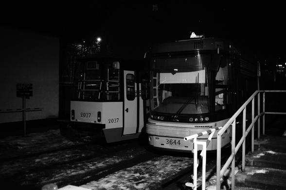 A high-contrast, black-and-white nighttime photograph of two trams parked side-by-side on snow-covered tracks. The composition is a medium shot, focusing on the front ends of the vehicles.
On the left, an older, more angular tram model (numbered 2037) features a boxy design with a flat windshield and vertical lines. To its right, a modern tram (numbered 3644) displays a more aerodynamic, curved front profile with a large, wraparound glass windshield. The lighting is harsh and directional, likely from overhead depot lamps, creating deep, rich blacks in the shadows and bright, reflective whites on the snowy ground and the trams' metallic surfaces.
The textures of the slushy snow and the metal railings in the bottom right foreground are sharply defined. The background is almost entirely enveloped in dark shadow, emphasizing the industrial silhouettes of the vehicles. The image has a slight grain and a distinct tonal depth that suggests it was shot on a vintage lens, specifically the Carl Zeiss 35mm f/2.4 Jena Flektogon mentioned in the post, which provides a wide, immersive field of view with characteristic sharpness and micro-contrast. The absence of digital noise in the shadows and the specific blooming around bright highlights suggest this may have been shot on high-contrast monochrome film or a high-end digital sensor mimicking a cinematic film aesthetic.