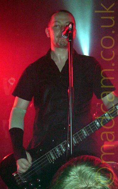 A man with close-cropped dark hair, wearing a black shirt and with his elbows and wrists strapped up, playing a black bass guitar and standing, grimacing, behind a microphone on a stand.
Red light floods the background, with a single white light shining upwards behind his left shoulder.
The head of a fan watching the gig is right at the bottom of the frame.