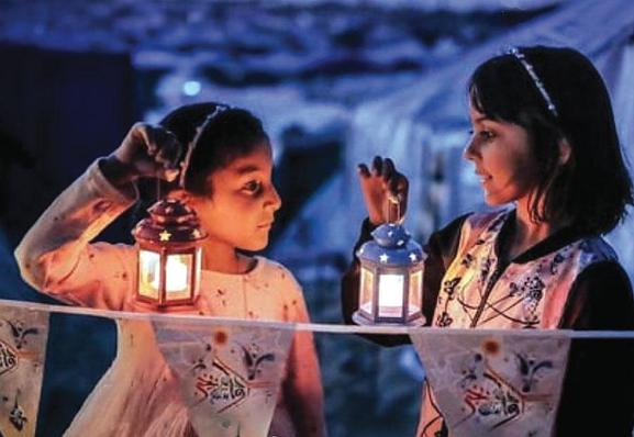 Photo. Derrière des fanions, deux petites filles se faisant face sont éclairées par les lanternes qu'elles tiennent. En arrière-plan, dans un flou de couleur bleue, on peut deviner les nombreux abris de fortune.