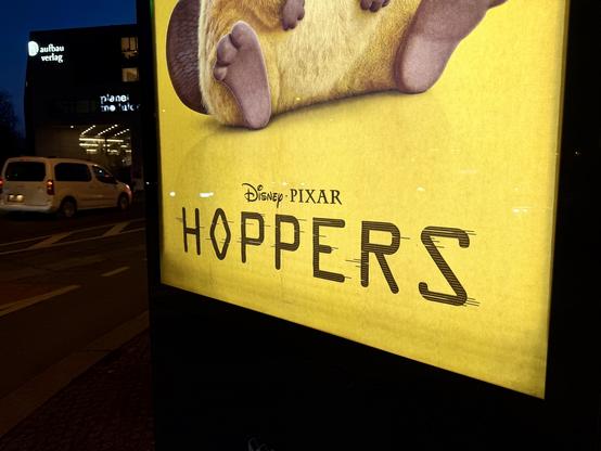 Lower part of a all-yellow movie poster for the film Hoppers placed at road where a car passes and a building is the faint background; the poster shows the logos of Disney and Pixar with a cropped lower body of a furry cartoon animal sitting