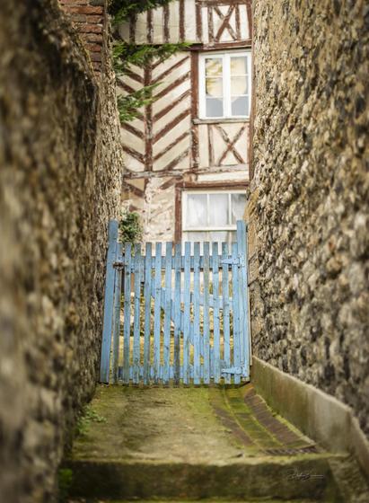 A narrow pathway between two stone walls leads to a blue wooden gate. In the background, a half-timbered house with a window is visible. The scene is atmospheric and evokes a sense of hidden charm.