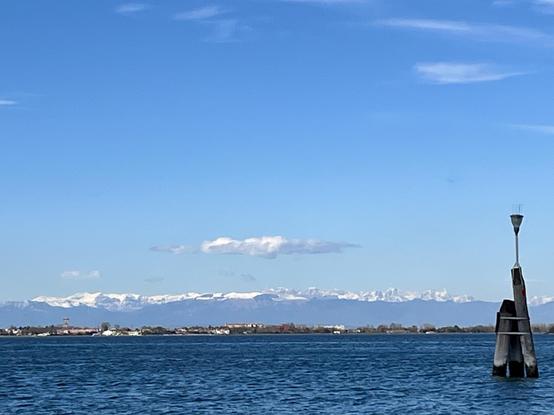 Looking out over the Venetian lagoon on an astonishingly clear day. In the foreground on the right, a traditional three legged navigation post made of dark grey wood, with something that looks like an upturned sink plunger attached to the top of it, which I think is meant to deter perching birds. The lagoon water is an unusual shade of navy blue, and gently rippled. The horizon line is about a quarter of the way up the frame, and is a view of an island with low houses and trees. Behind the island in the far distance, but very clearly visible, the snow capped peaks of the Dolomites rise into the vast blue cloud-flecked sky.