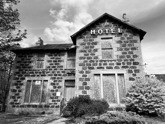 An old, abandoned hotel building made of stone, with boarded-up windows and a "HOTEL" sign on the front. The image is in black and white, featuring overgrown bushes and a cloudy sky.