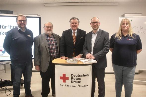 Vier Personen stehen vor einem weißen Tisch mit Broschüren. Logo des Deutschen Roten Kreuzes ist erkennbar. Heller Raum.