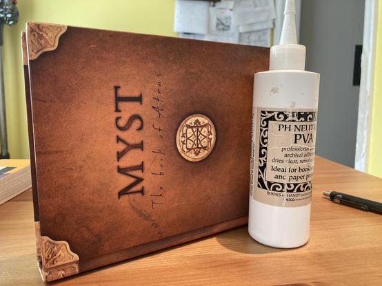 A hardcover copy of Myst: The Book of Atrus by Rand Miller, Robyn Miller, and David Wingrove. It is resting on its spine. A large bottle of PH Neutral Polyvinyl Acetate (PVA) glue sits beside it upright, holding the book upright.