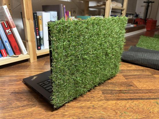 A laptop with its lid covered with fake grass. It’s sat on the floor in front of a bookshelf, with some fake grass scattered around it