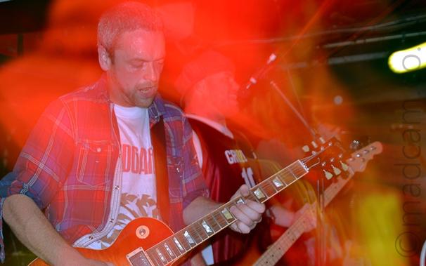A man with short, close-cropped greying hair and beard, and wearing an open blue, red, and white checked shirt over a white t-shirt, playing an orange electric guitar.
A camera flash has frozen him in place, while a long exposure and a slow zoom in has combined with the stage lights to paint the area around him with red and yellow light.
In the background, a man wearing a blue cap and wearing a red and white basketball shirt  and playing a bass guitar is singing into a microphone.
Black walls mark the edges of the room, with a bright light smeared into a streak in the top right corner.
