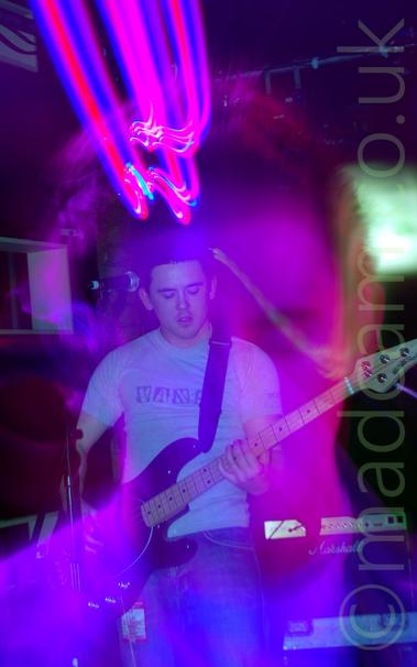 A man with short dark hair, wearing a grey t-shirt with a black and grey "Vans" logo across the chest, playing a black bass guitar.
An initial camera flash has frozen him in place, while a long exposure and a snap zoom has created a somewhat soft close-up of his face, illuminated by purple stage lights.
The whole image has a purple cast, with streaks of purple lights at the top.