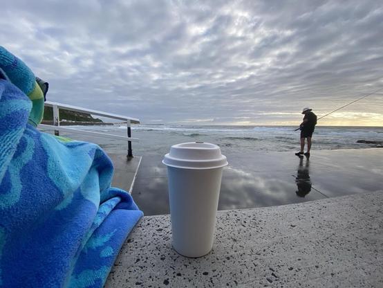 Tall white takeaway coffee cup sitting on a concrete step overlooking the water at the Cowrie Hole, looking northwards towards Nobbys Head. There is a blue and green patterned beach towel to the left. The fisherbloke is standing to the right on the edge of a wide wet pathway. The ocean runs next to that edge, occasionally splashing up - thus the wet path. Cloudy day with glimpses of morning light. Beautiful cloud reflections on the path almost look like steam coming out the sides of the coffee cup.