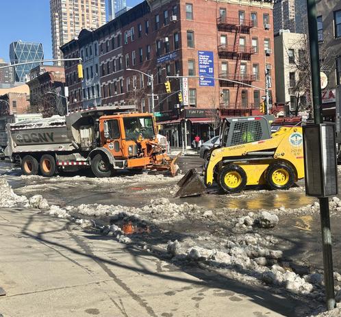 Brightly colored trucks move snow, NYC midtown buildings, some high rises in background.