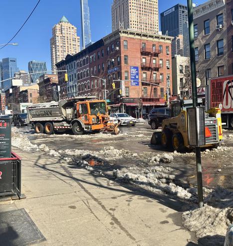 Orange dump truck being filled with snow. NYC midtown buildings, some high rises in background.