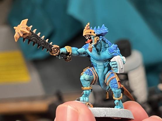 A miniature of a Saurus Warrior from Age of Sigmar. The bipedal man sized lizard has blue scales, while being adorned with golden jewelry and regalia. The golden mask resting on its face seems to give some indication of it leading a unit. In its right hand is a massive spiked club while the other hand has a glue point for a shield.
