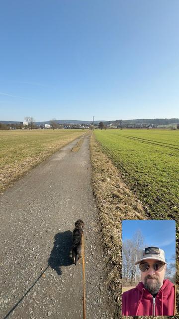A dog walking on a gravel path through a field under a clear blue sky. Inset photo of a person wearing sunglasses and a cap, smiling.