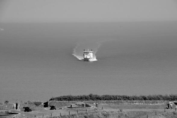 This black-and-white photograph captures a ferry or passenger ship traveling across a body of water, leaving a prominent wake behind it. The ship is centered in the image, moving away from the viewer toward the horizon. The sea appears calm, with gentle waves visible.
In the foreground, there is a grassy coastal area with a few small structures, possibly old military bunkers or lookout posts, and a couple of people standing near the edge of the land. The horizon is faintly visible, and the overall atmosphere of the image is serene and nostalgic. The sky and water blend into a uniform gray, emphasizing the timeless quality of the scene.