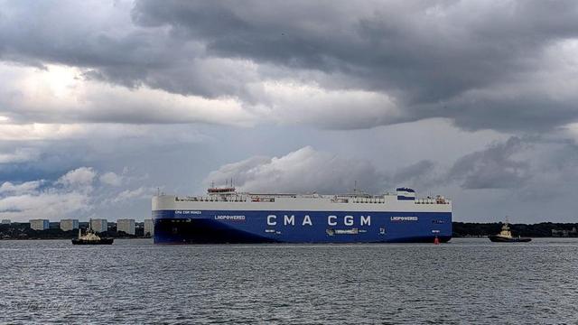 The image shows a large cargo ship named "CMA CGM Monza" sailing on a body of water. The ship is predominantly blue and white, with the letters "CMA CGM" prominently displayed in large white letters on the side of the hull. The words "LNG POWERED" are also visible, indicating that the ship is powered by liquefied natural gas.
The ship is accompanied by two smaller tugboats, one on each side, assisting it as it navigates through the water. The sky above is filled with dramatic, dark clouds, suggesting an overcast or stormy atmosphere. In the background, a city skyline with multiple buildings is visible along the horizon, indicating that the ship is near a port or coastal city. The water is relatively calm, with gentle waves.