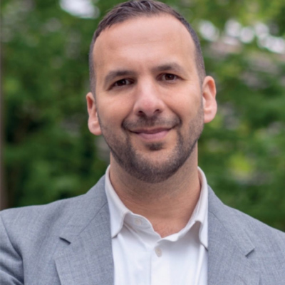 Headshot of Zack Polanski.  Grey jacket, white open necked shirt. Foiliage out of focus in the distance offering a green backdrop.