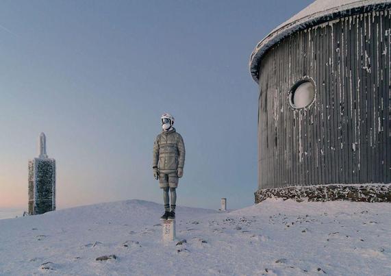 A figure in a padded jacket and helmet stands on a snowy landscape, near an observation building and a tall stone marker.