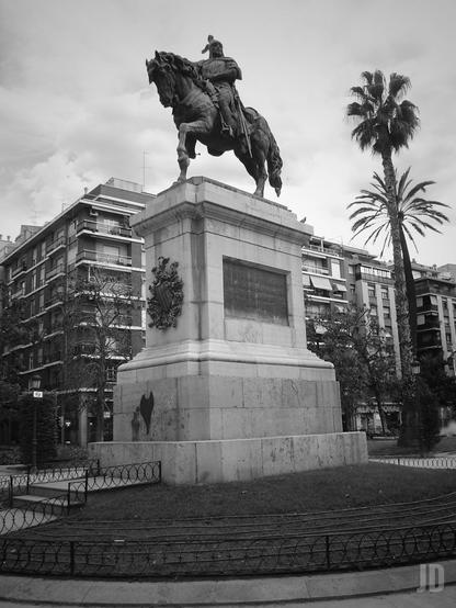 La imagen muestra una estatua ecuestre situada en el centro de una plaza urbana. La escultura representa a un jinete con armadura montado sobre un caballo, ambos sobre un pedestal de piedra rectangular con inscripciones y un escudo decorativo. Alrededor se ven edificios residenciales, palmeras y una zona ajardinada con barandillas bajas. El cielo está nublado y la fotografía está en blanco y negro.