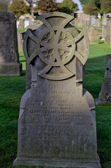 Photo format portrait montrant une pierre tombale ancienne avec une croix aux 4 branches identiques, reliées par un cercle. D’autre tombes sont visibles en fond