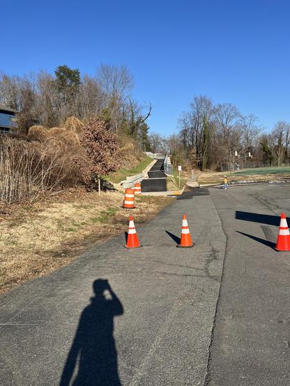 Color photo showing a parking area behind Orange safety cones, then a new asphalt path up a steep hill