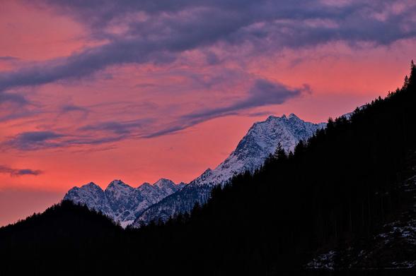 Beschneite Berge vor morgenrotem Himmel. Der rötliche Himmel ist mit Wolken durchzogen, von links unten ist eine kleine baumbewachsene Erhebung in Pyramidenform die dann fließend in einen stetig nach rechts oben steigenden Hang übergeht. Ab etwa der Pyramidenspitze und oberhalb des ansteigenden Waldes sind im Hintergrund diese beschneiten Berge zu sehen.