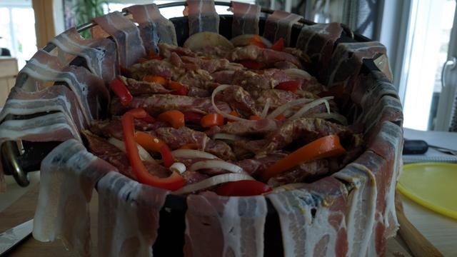 ein Dutch Overn, Speck hängt heraus und es ist bereits Fleisch, Zwiebeln und Paprika eingeschichtet