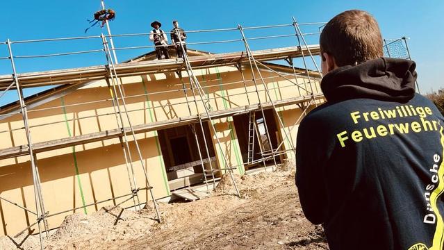 Baugerüst an einem hellgelben Haus. Zwei Personen arbeiten auf dem Dach. Im Hintergrund blauer Himmel. Eine Person mit schwarzer Jacke mit Aufschrift "Freiwillige Feuerwehr" im Vordergrund.