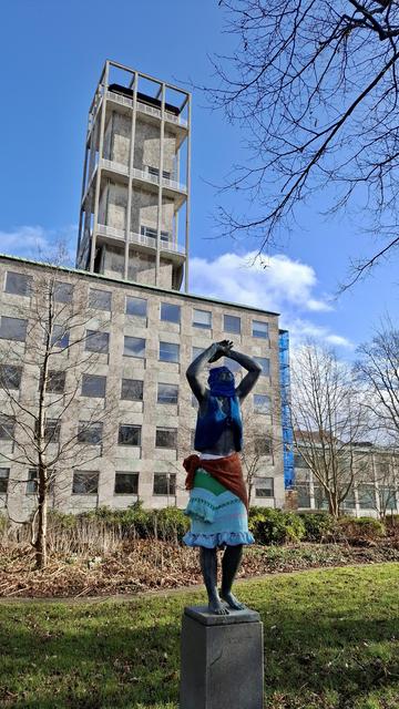 Short version: statue of woman in knitted clothes. Fuller description: The statue 'Vaagnende kvinde' ('Waking woman') erected in 1920, on a plinth at Rådhuset, Aarhus City Hall. A figure of a woman on a plinth. Sunshine and shadow and deep blue sky with white clouds above. The rectangular City Hall clock tower rising behind her. The life-size figure has one leg slightly raised, as if taking a step, with arms raised above her head. Someone has dressed her in a turquoise and green skirt; a dark orange sarong wrap; a dark blue bolero; and a dark blue head covering. All knitted and lovely.