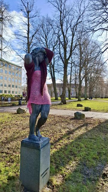Short version: statue 'Girl' in knitted clothes. Fuller description: the statue Pige ('Girl'), from 1940, on a plinth at Rådhuset, Aarhus City Hall, Denmark. A figure of a  young woman on a plinth. Sunshine and shadow and deep blue sky with white clouds above. Trees and a corner of city hall beyond. The life-size figure has one leg slightly raised, as if taking a step, with arms folded around her head, the head to one side. As if sad or afraid or wary. Someone has dressed her in a bright light pink strapless dress and wrapped a dark purple scarf around her arms and head. All knitted and lovely.