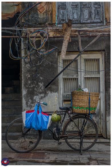 a bicycle, with a blue bag, most likely filled with straws and few other items, then in between you see two tender coconuts tied around, the basket on the back is also full of coconuts. 

This is basically a person who uses this to sell coconut water to people on the street. 

Behind it is a wall with black and white (now grey) and a window with metal grills on them (closed) - on the left you see stairs taking you upstairs. the floor upstairs has the doors bolted. 

between the two floors you see long pipes, wooden planks, and long folded thick wires for electricity.
