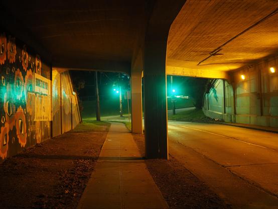 A traffic  underpass in the pre-dawn morning. There is a brightly colored mural on the wall on on pedestrian side illuminated by the green traffic lights at the end of the tunnel and yellow lights on the traffic side of the tunnel.
