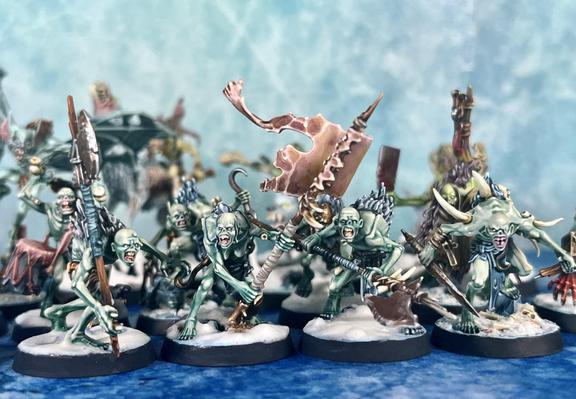 a bunch of light green skinned ghouls, very skinny and hungry, on snow & ice bases over a blue ice mat, wielding halberds, rusty knives and a battle standard made from human skin. There are more (blurry) monsters in the background.