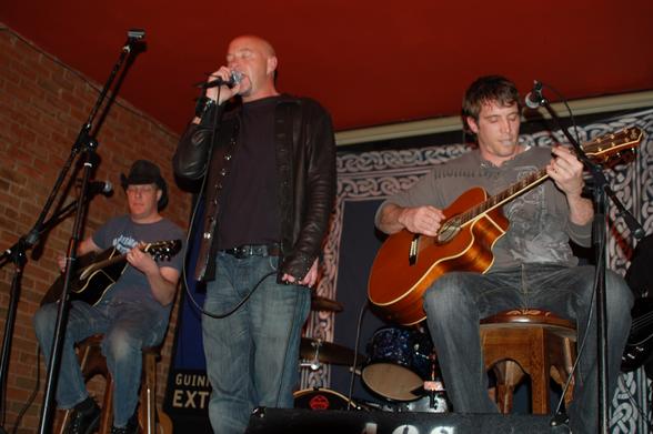3 members of a band on a stage.
On the left is a man wearing a black stetson, a grey t-shirt with a white logo, and a blue pair of jeans, sitting on a stool and playing a black acoustic guitar.
In the middle is a man with a shaven head and a thin goatee beard, wearing a leather jacket over a black t-shirt and a blue pair of jeans.
He is holding a microphone in his right hand, close to his mouth as he sings in to it, holding the cord in his left hand down by his waist, with his eyes closed.
On the right is a man with dark hair, wearing a grey, long-sleeved t-shirt and grey jeans, sitting on a wooden stool and playing a brown acoustic guitar, with a guitar pick held in his lips.
A blue cloth with a white knotted design around the edges covers the wall behind them, with a red brick wall on the left, with a red ceiling above them.