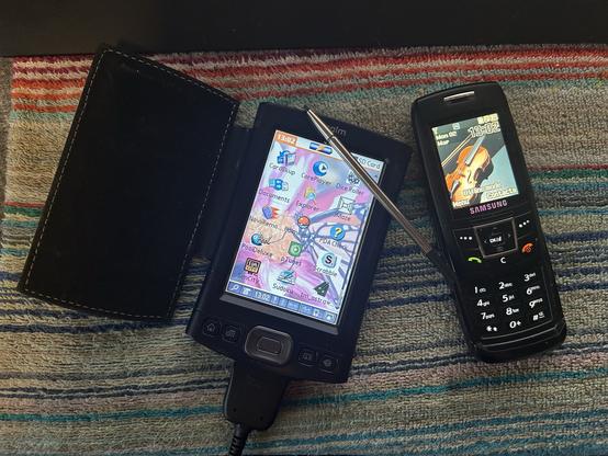 A Palm TX showing the application launcher and a Samsung E350 slider mobile phone open on a desk.