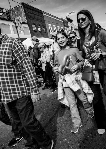 A black and white Dutch angle photo of a young girl with a twisted balloon.  There are other people all around due to a street festival going on.