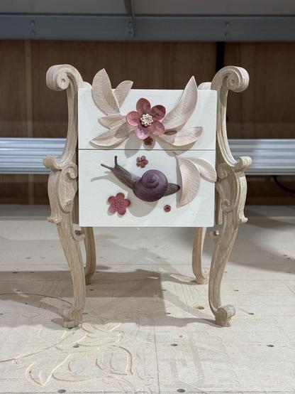 A pale plywood cube box with ornate legs and a snail and a flower for drawer knobs. There are leaves and flowers carved in wood as decorative element.