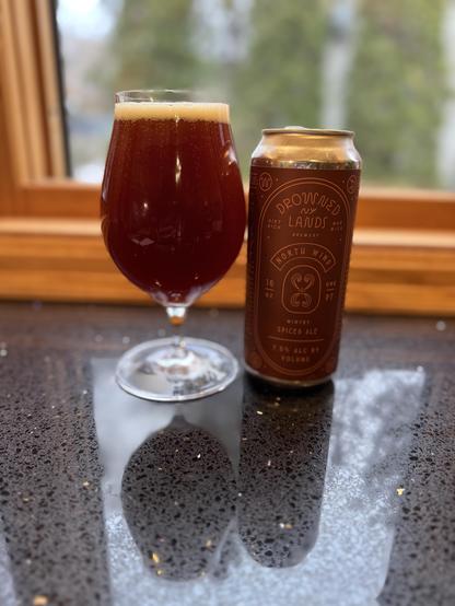 A tall snifter glass full of ruby colored ale is next to a similarly colored pint can on a shiny black countertop next to a window