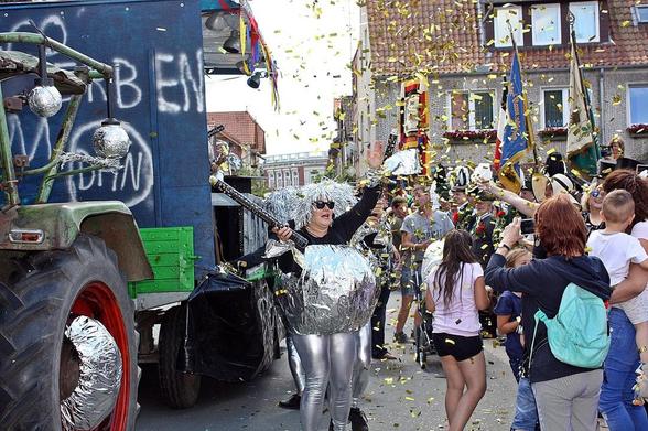 Straßenfest mit bunt kostümierter Person, die mit einem Besen tanzt. Im Hintergrund ein blau-grüner Lastwagen und jubelnde Menschen. Konfetti liegt auf der Straße.