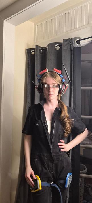 this one standing in front of a curtain wearing its ar glasses, headphones, and a set of black coveralls. the covetalls are unzipped, and a bit of its bra is visible. it is looking straight at the camera such that one eye is visible while the other is hidden under a refluction from the ar glasses' prisms. one hand rests on one of its keyboards