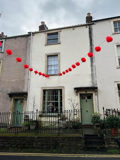 Two strings of bright red, decorative paper lanterns are strung across a street, presumably to celebrate the Chinese new year. The lines are anchored to the first-storey window ledge of a mid-terrace townhouse. The building is built on a slope, opens directly to a footpath and faces an iron railing. The rendered facade is a magnolia colour and the doors and window frames have been painted laurel green, and the windows (one per floor) reduce in size and complexity as they rise up the building (20 panes, 12 panes, 6 panes). Phoenix Terrace, Catherine Street, Frome, Somerset, UK. Photo by Gearóid Burke.