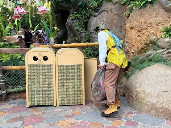 A Tokyo DisneySea cast member carries replaces a trash bag toward themed trash cans nestled into rockwork and oversized foliage near Tinker Bell’s Busy Buggies in Fantasy Springs.