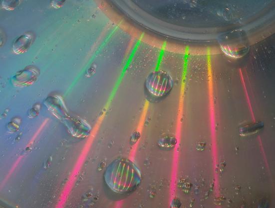 A wet CD was illuminated outdoors during the day with white LED light. The CD's surface refracts the light rays, which are then reflected and refracted by the droplets. The center of the CD, of which only a small portion is visible, appears like a dark sun from which thin beams of light emanate, each in several rainbow colours. These beams of light are visible within the droplets.