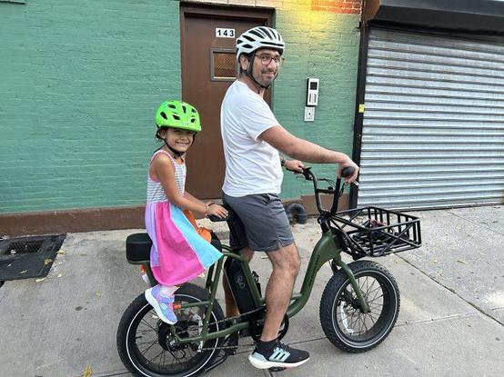 Adult and young child wearing bike helmets ride together on a cargo e-bike on a city sidewalk, the child seated behind the adult and smiling, suggesting everyday family life and climate-friendly urban mobility.