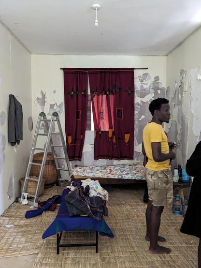 A man in a yellow T-shirt and camouflage shorts stands barefoot in a small room with peeling paint on the walls. A metal ladder stands near a woven basket. A low bed with patterned bedding sits beneath red curtains covering a window. In the foreground, folded clothes rest on a small table over a woven mat floor, while household items and a lantern sit on a side surface to the right.